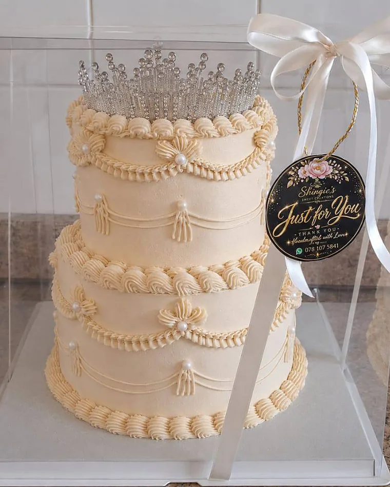 Two-tier ivory celebration cake with piped pearl detailing and a crystal tiara, boxed and ribboned