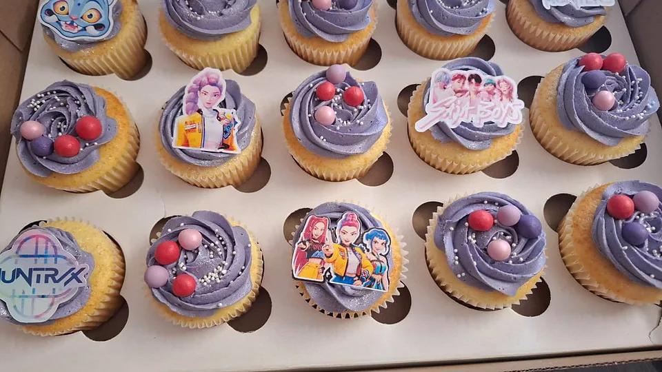A box of themed cupcakes with lilac swirls and printed character toppers